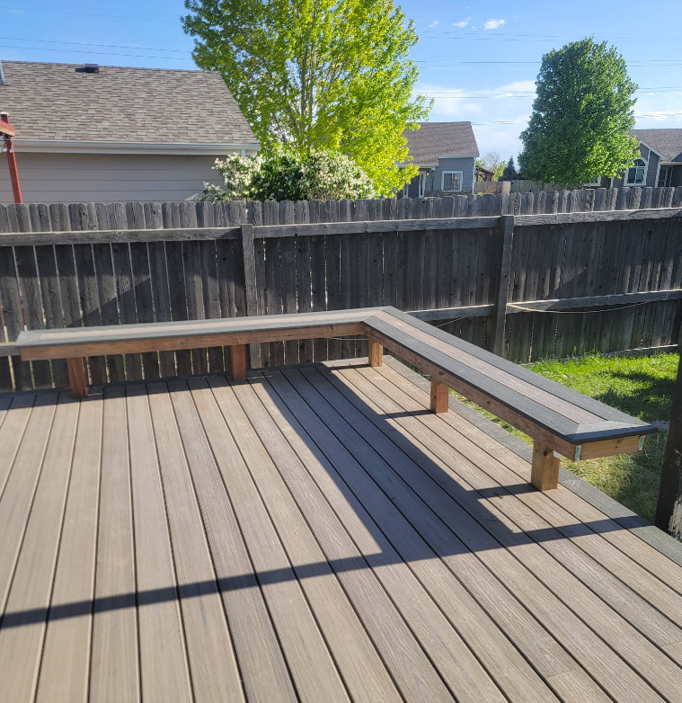 L-shaped wooden bench on a tan deck in a backyard with a wooden fence and trees in the background.