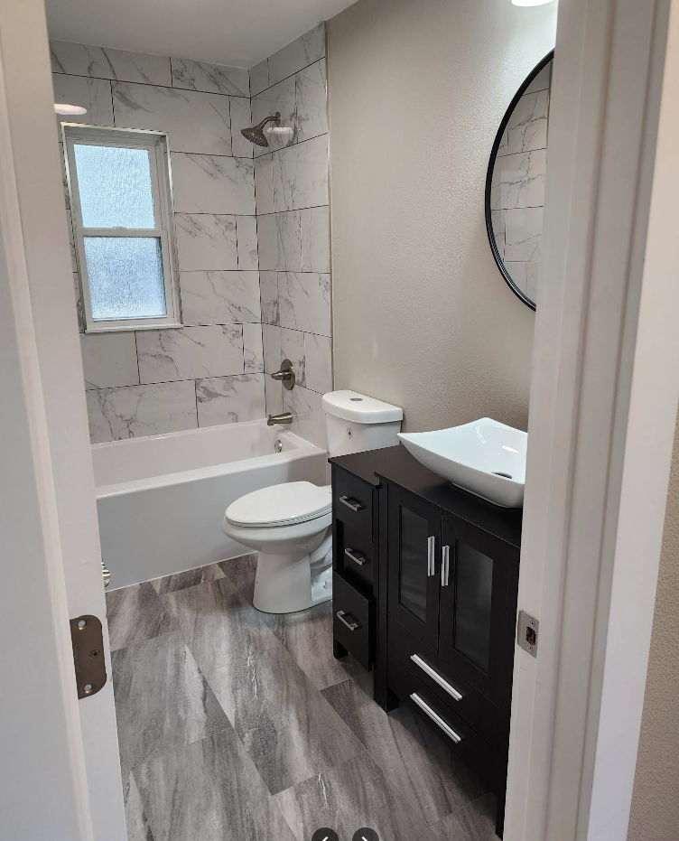 A modern bathroom featuring a marbled tiled tub, a white toilet, and a dark wooden vanity with a square vessel sink.