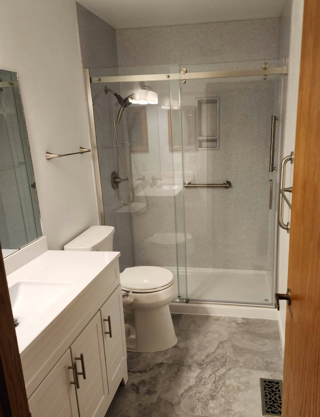 A modern bathroom featuring a white vanity, a toilet, and a glass-enclosed shower with gray tiled walls and flooring.