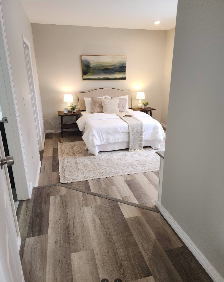 A bedroom view from a hallway, featuring a bed with white linens, two side tables with lamps, and wood-look flooring.