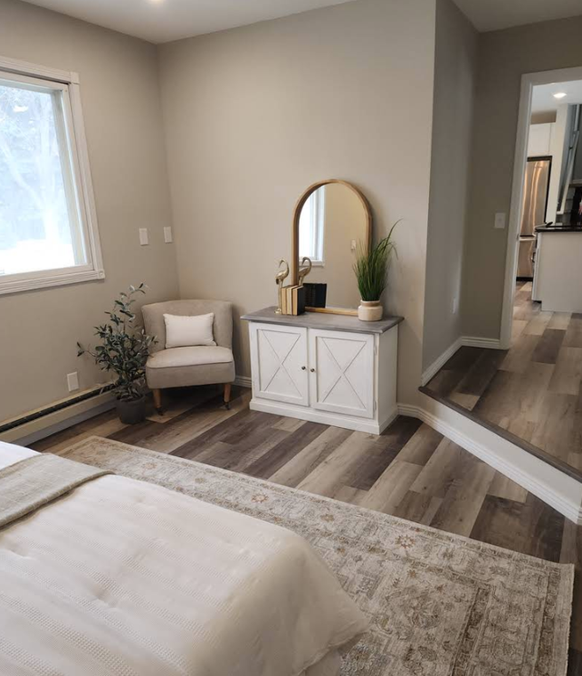 A cozy bedroom corner with a white cabinet, arched mirror, armchair, potted plant, and a textured rug on wood flooring.