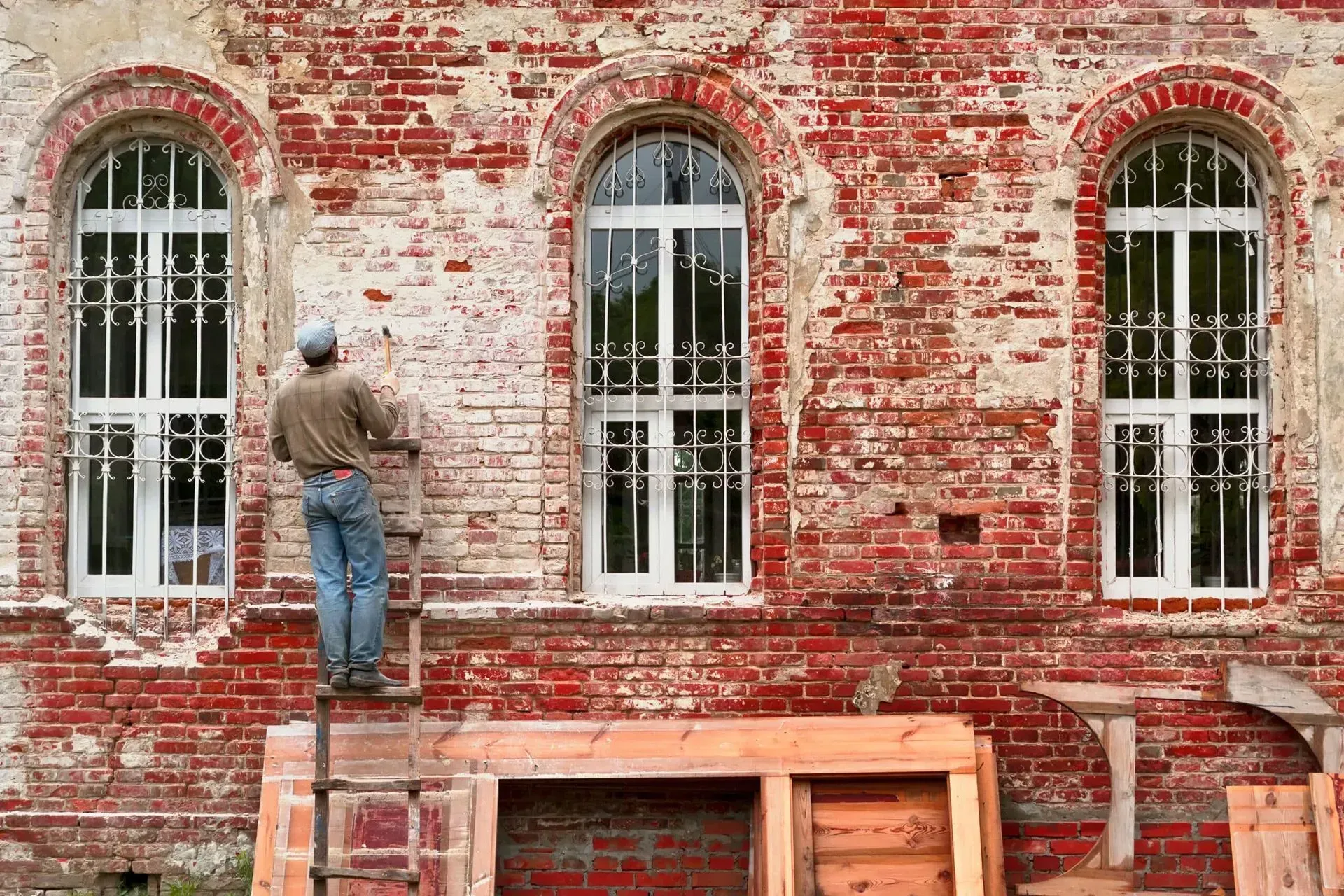 A person stands on a ladder, working on the red brick wall of a building with three arched windows.