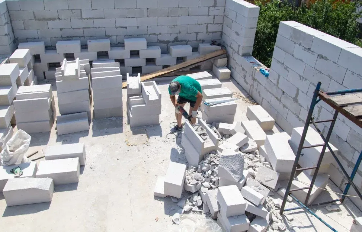 A person in a green shirt works with a power saw on white building blocks at an active construction site.