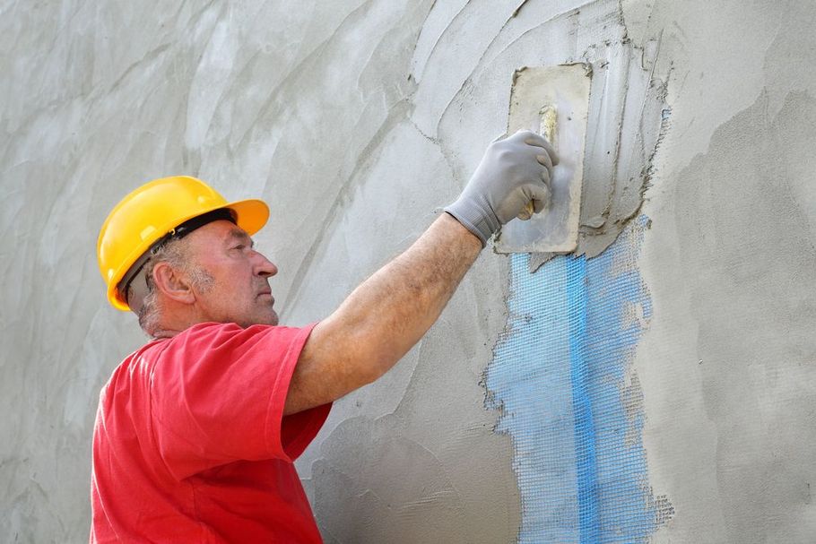 A construction worker in a red shirt and yellow hard hat uses a trowel to apply plaster over blue mesh on a gray wall.