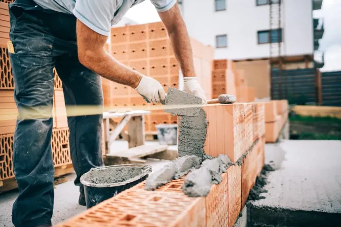 A construction worker in work pants and gloves applies mortar to red ceramic bricks on a construction site.