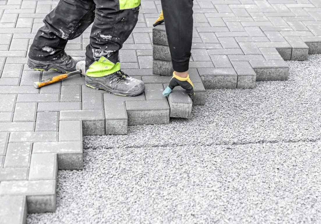A construction worker wearing safety gear places gray interlocking paving stones on a gravel base.