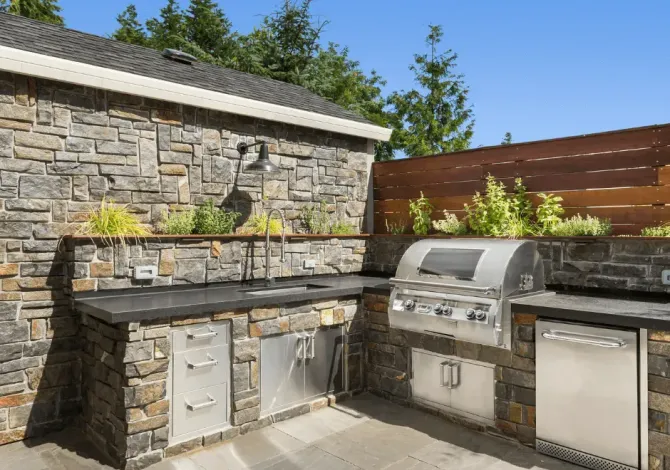 An outdoor kitchen featuring stone masonry, stainless steel appliances, a grill, and a built-in sink on a patio.