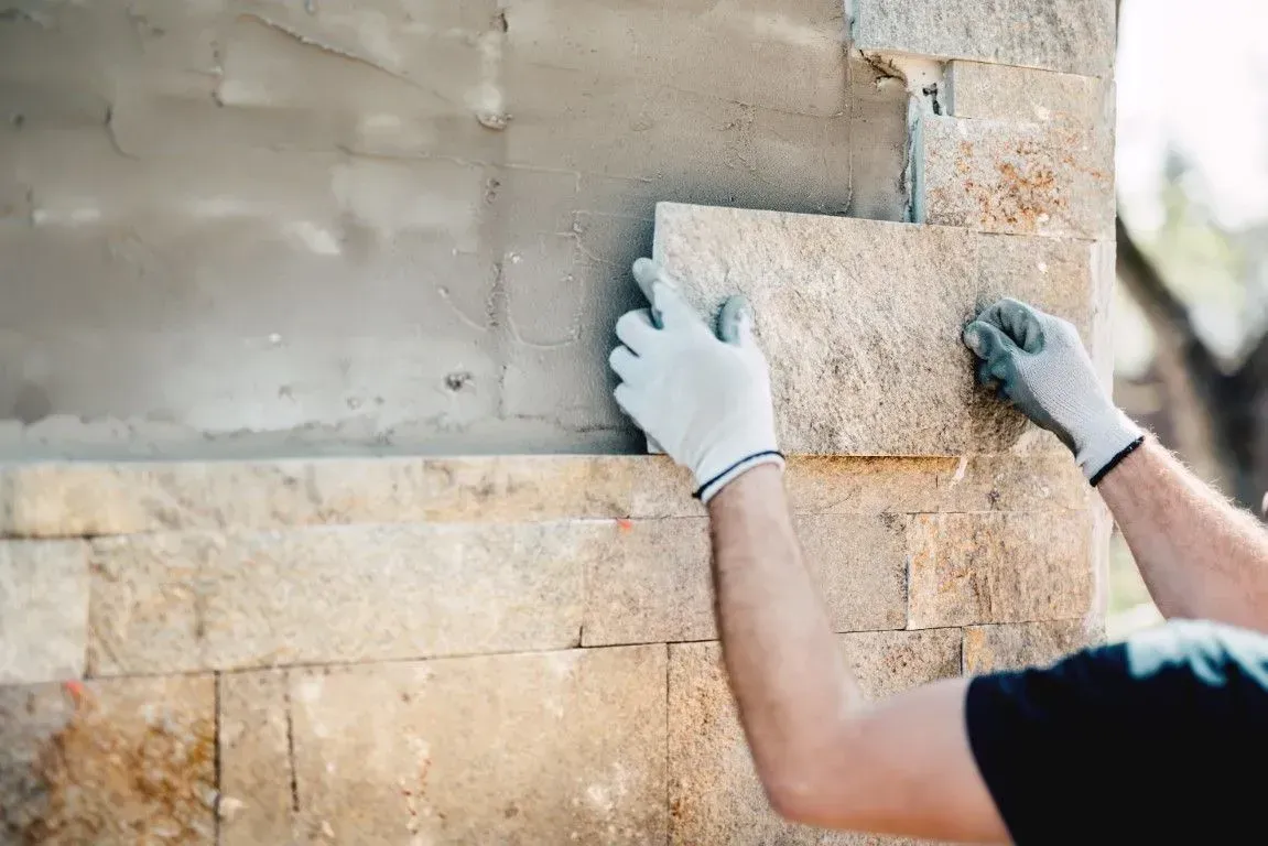 A person wearing work gloves places a rectangular stone tile onto a wall covered in adhesive mortar.