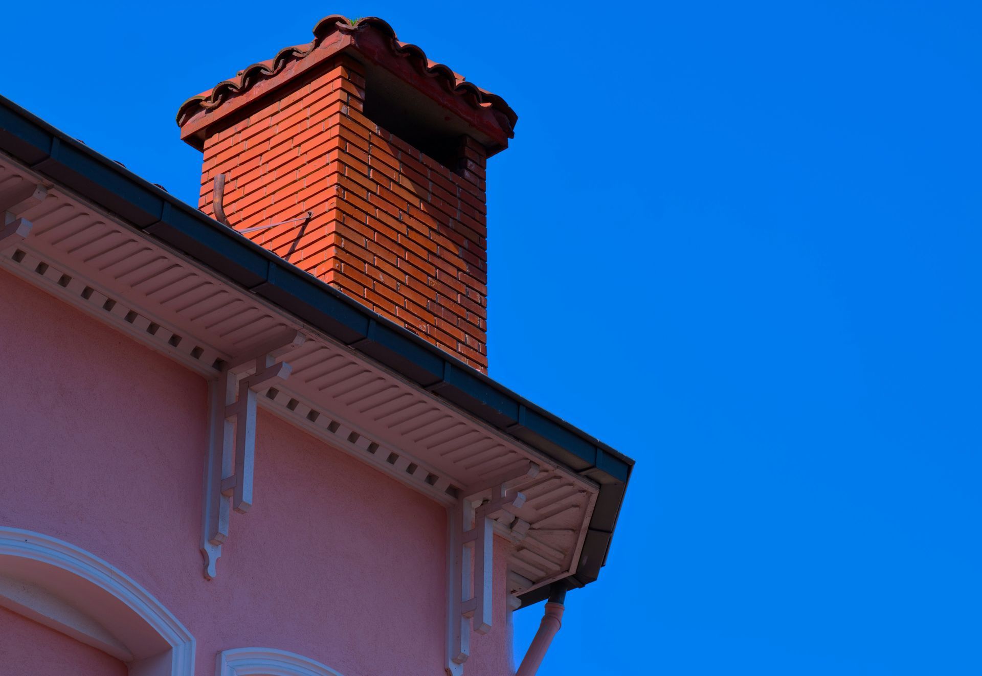 A red brick chimney rises from the edge of a light pink house roof against a vibrant, clear blue sky.