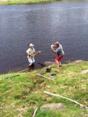 Two men are standing next to a river playing guitars.