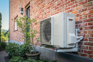 Outdoor HVAC unit beside a brick house wall, with a small tree and shrubs in the yard.