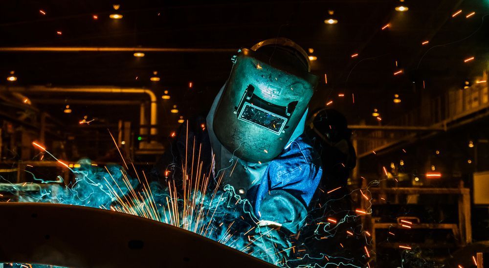 A man wearing a welding mask is welding a piece of metal in a factory.