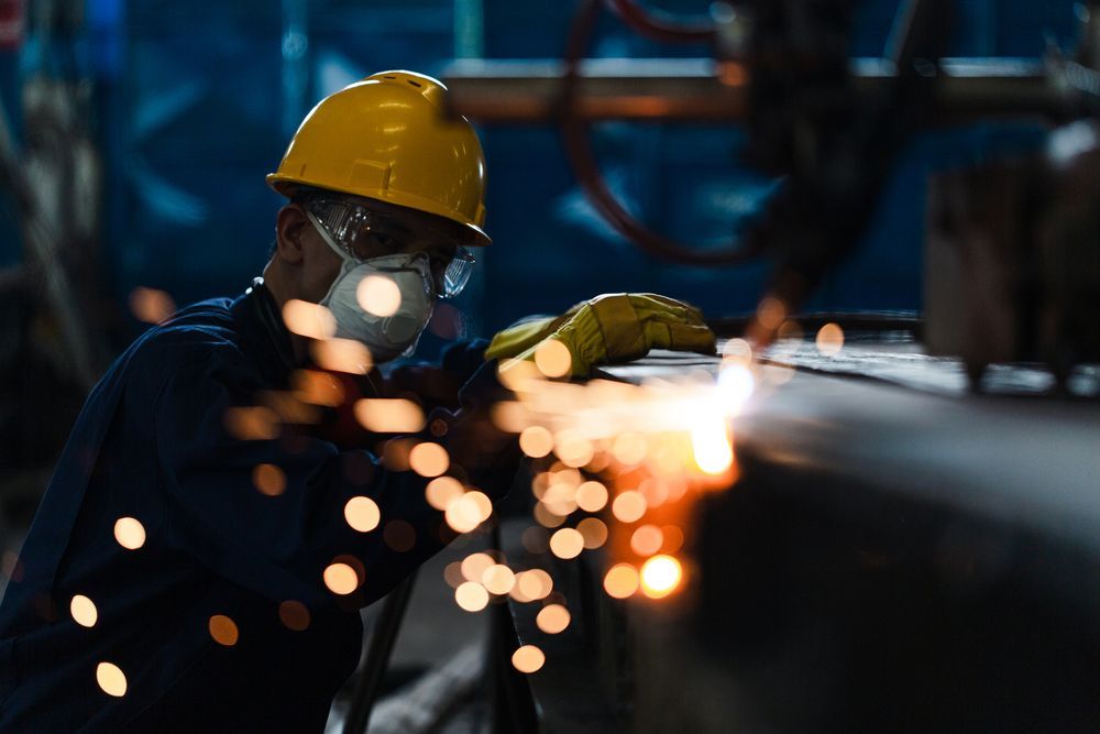 A man wearing a hard hat and a mask is welding a piece of metal in a factory.