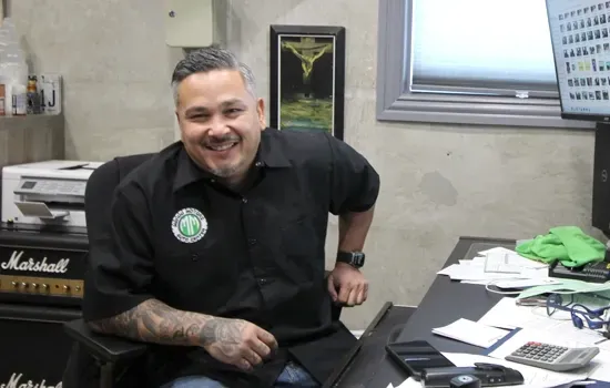 A smiling person with arm tattoos sits at a desk in an office with a Marshall amplifier and a framed picture on the wall.