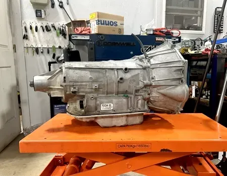 A metallic automotive transmission sits on an orange hydraulic lift table inside a garage.
