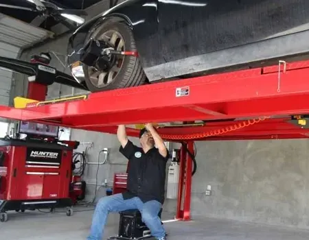 A mechanic works under a car raised on a red hydraulic lift inside a garage, with Hunter alignment equipment nearby.