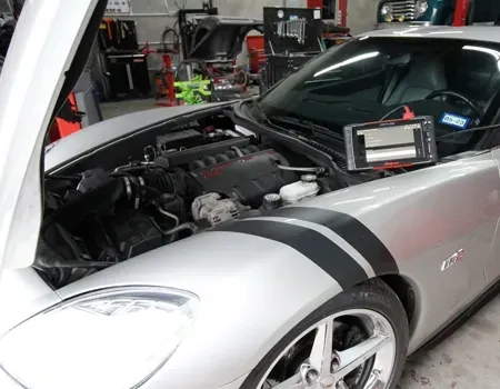 A silver sports car with an open hood is parked in a garage, with a diagnostic tablet resting on the windshield.