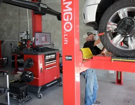 A technician in an auto shop works on a car tire lifted by a red hydraulic hoist next to a Hunter alignment console.