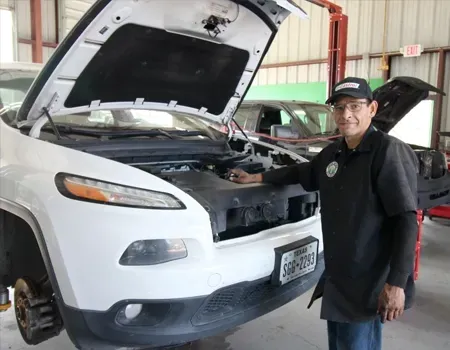 A technician in a black uniform stands next to a white SUV with its hood open in an auto repair shop.