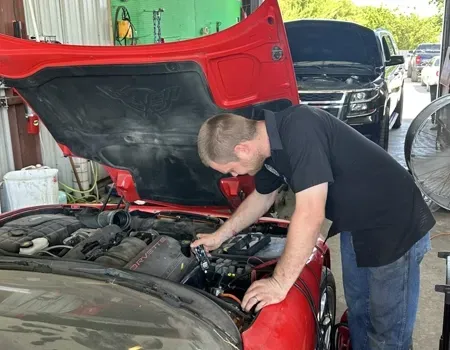 A mechanic leans into the open hood of a red car in a garage to inspect the engine.