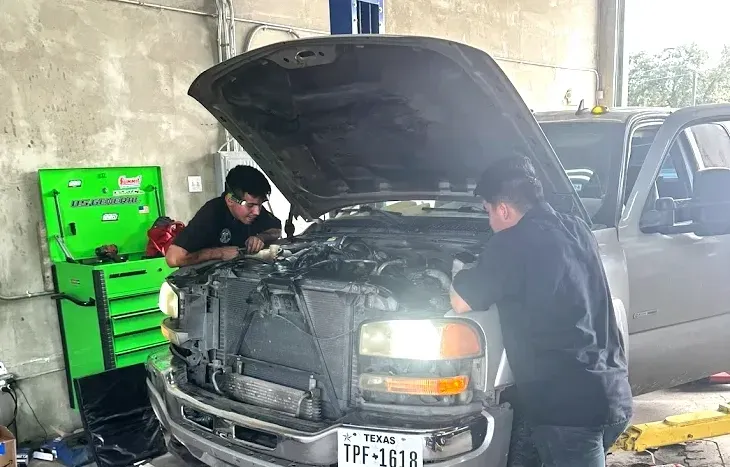 Two mechanics work on the engine of a silver truck with its hood open inside a repair shop with a green tool chest.