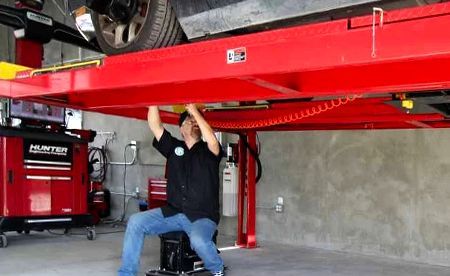 A technician sitting on a rolling stool works underneath a vehicle raised by a bright red hydraulic lift in a garage.