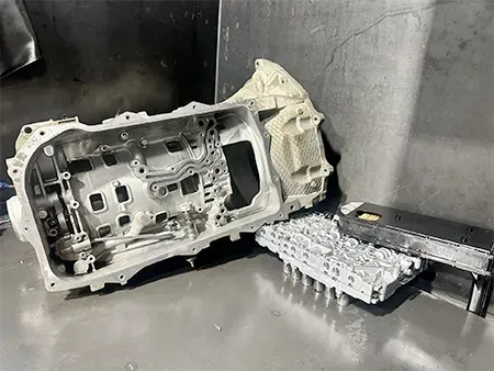 A metal transmission housing and its valve body component placed on a grey workshop surface.