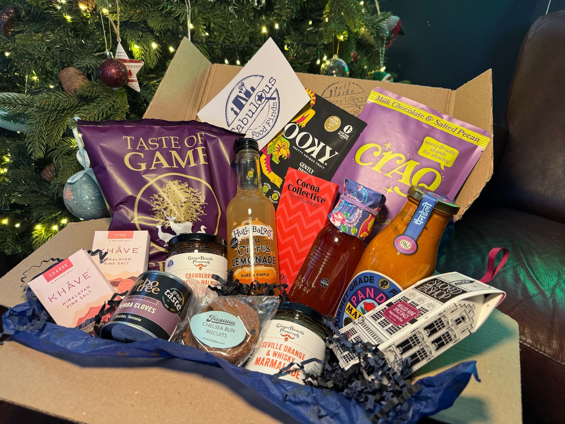 A box filled with food and drinks is sitting in front of a christmas tree.