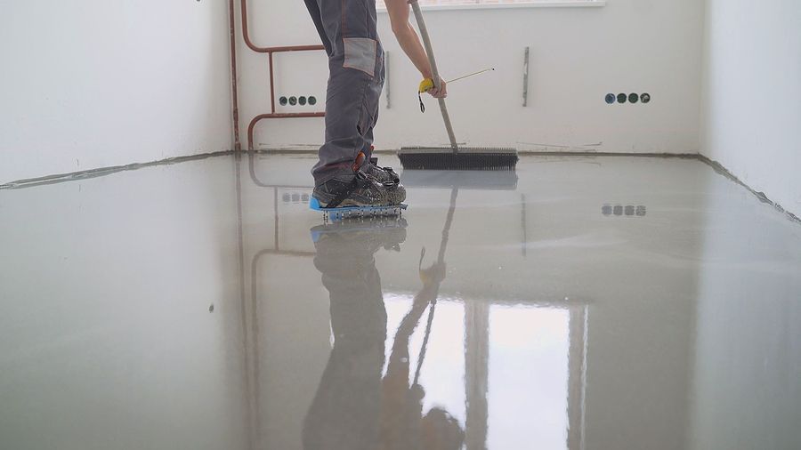 Person smoothing wet gray flooring with a brush while wearing special roller skates in a room.
