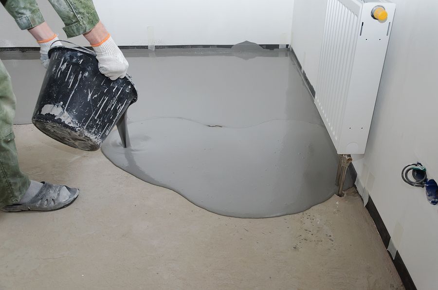 Person pouring gray concrete from a black bucket onto a floor near a white radiator.