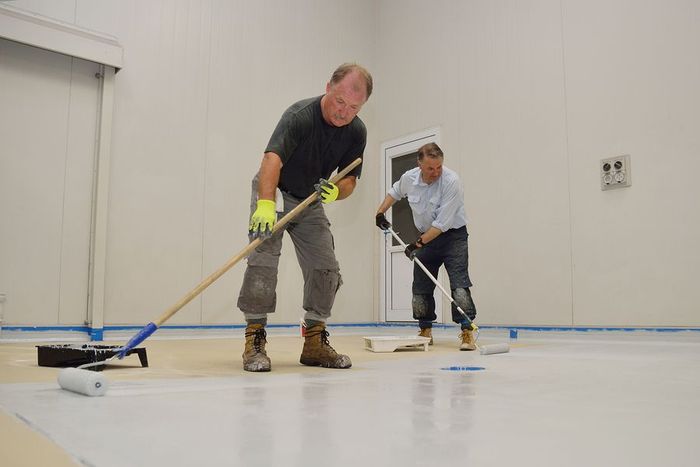Two men applying white coating to a floor with rollers in a bright, indoor setting.