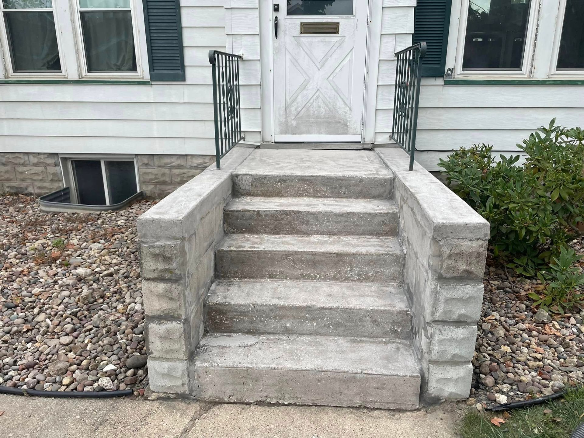 Concrete steps leading up to a white door, with black railing, flanked by gravel and a bush.