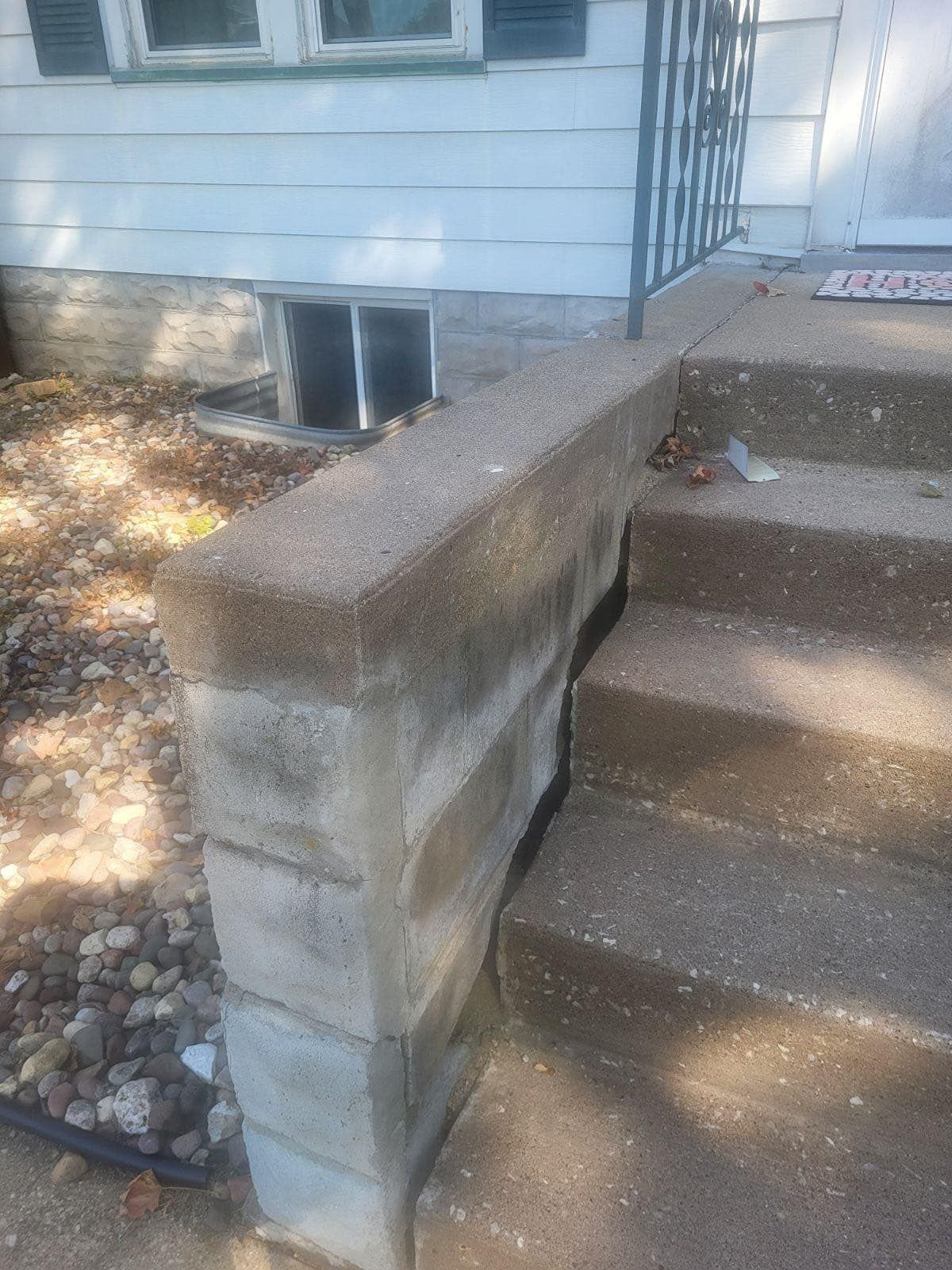 Concrete steps and retaining wall leading to a house entrance. The wall is made of stacked blocks, the steps and wall appear weathered.