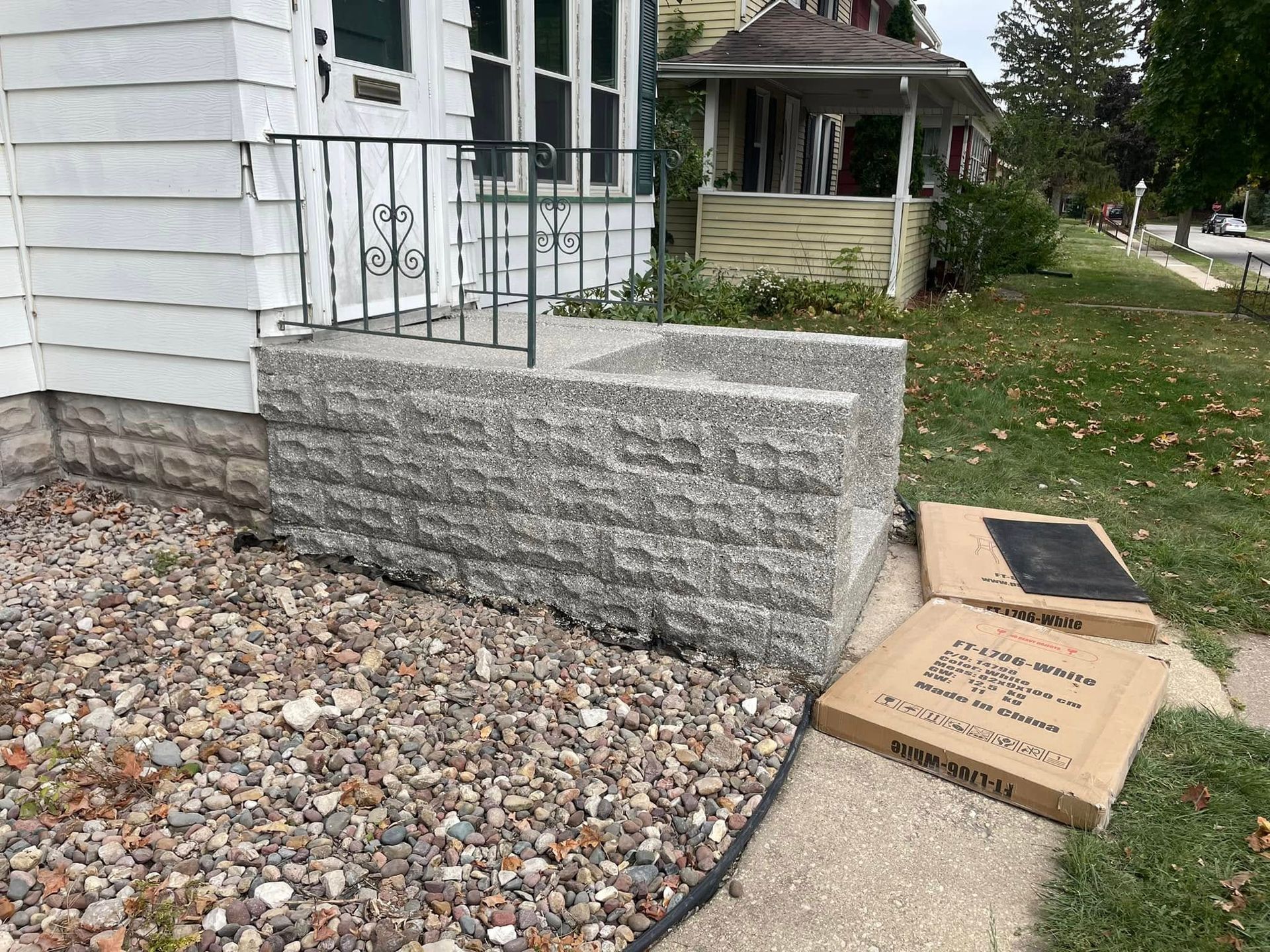 Gray brick porch with metal railing next to a white house; cardboard boxes on the sidewalk.