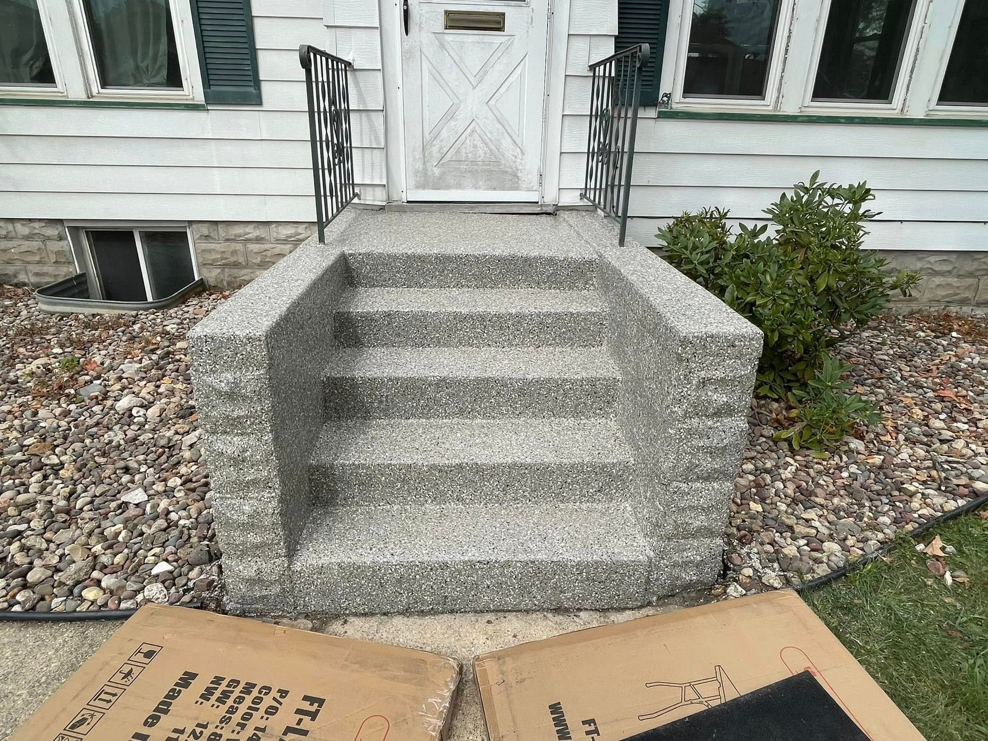 Concrete steps leading to a house entrance. Includes railing, door, and surrounding gravel. Two cardboard boxes in the foreground.