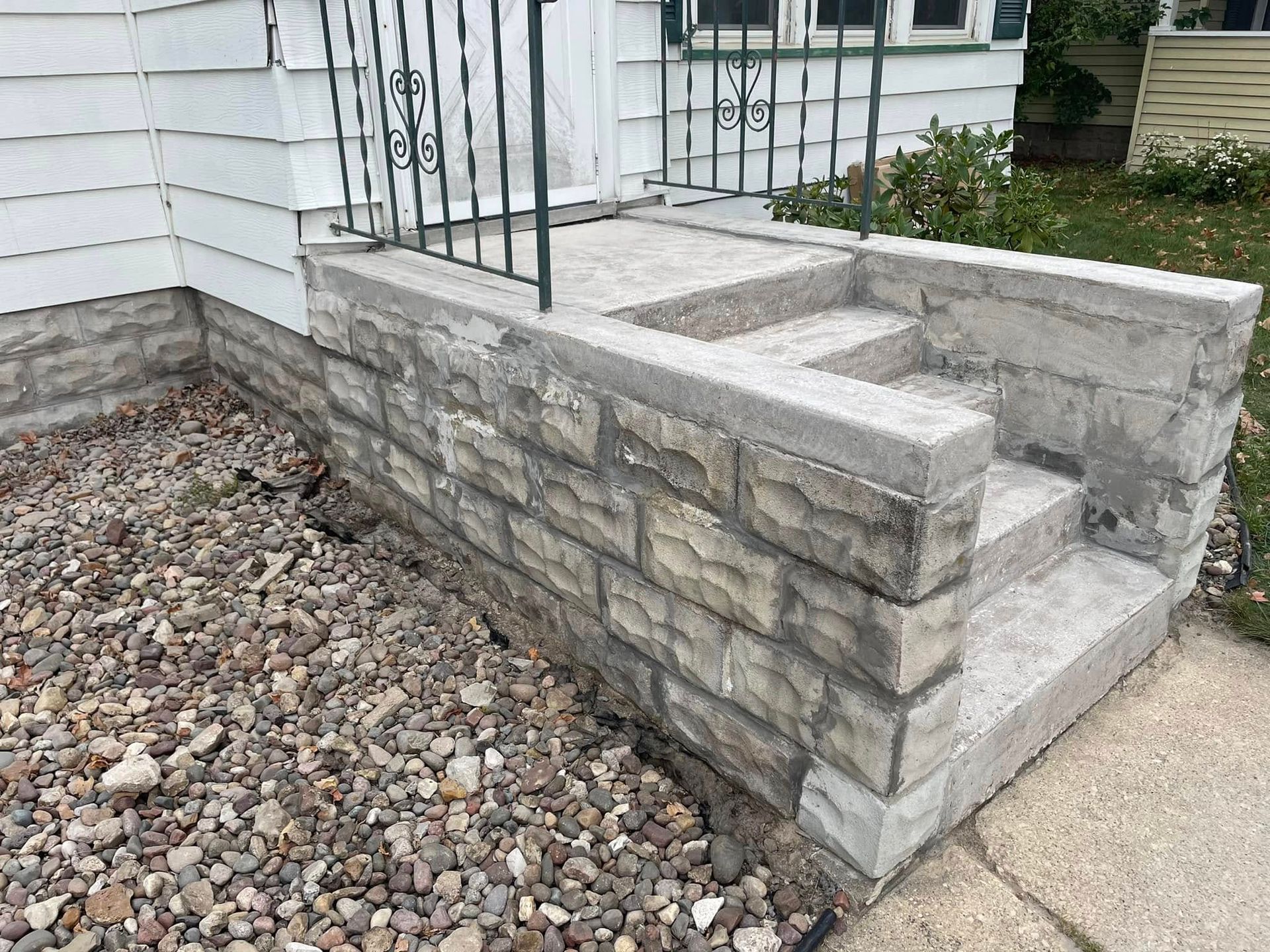 Stone steps leading to a white house with a dark railing. Gravel and sidewalk are visible.