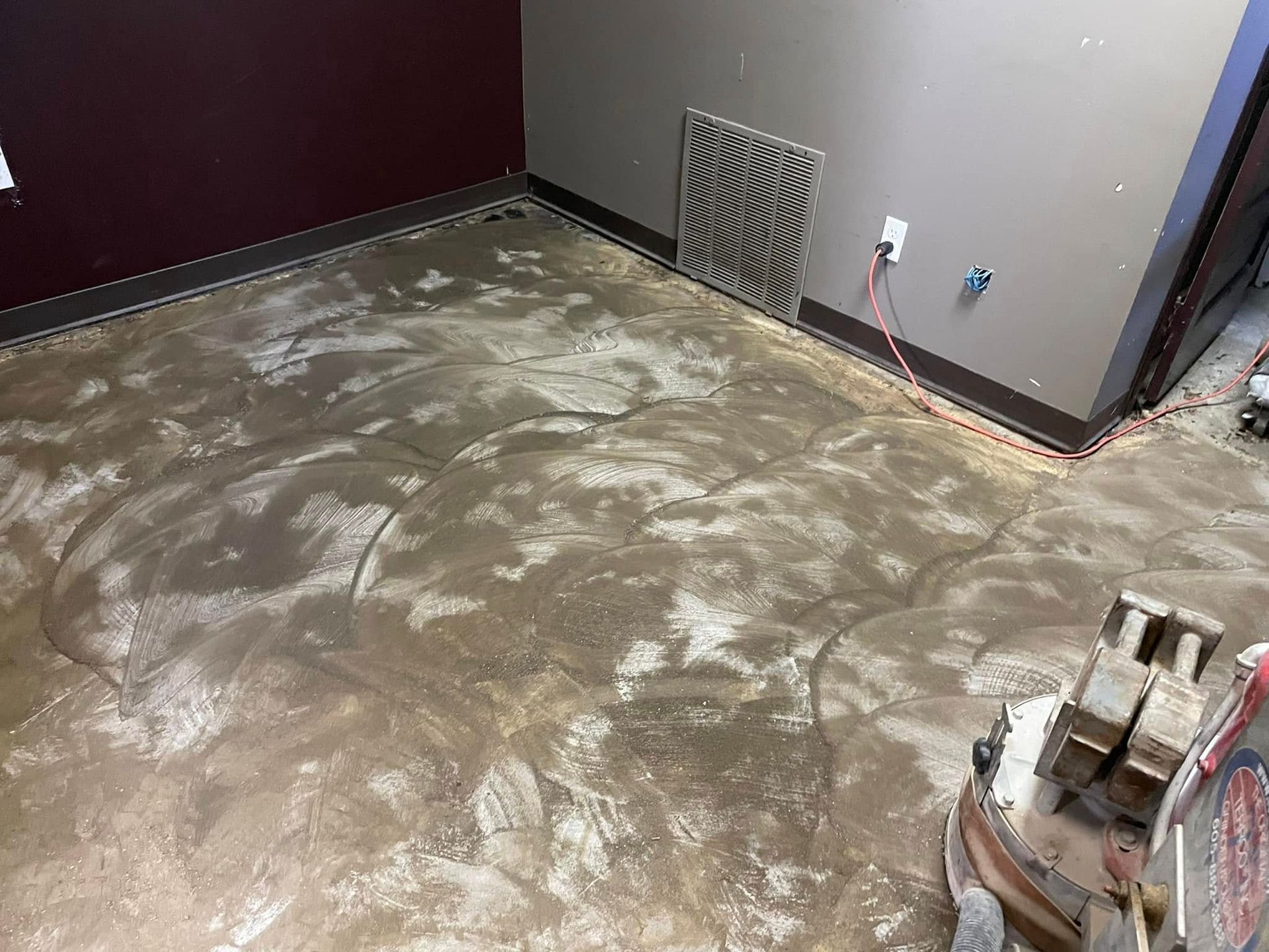 Concrete floor being ground, with visible swirling patterns, in a room with burgundy and grey walls.