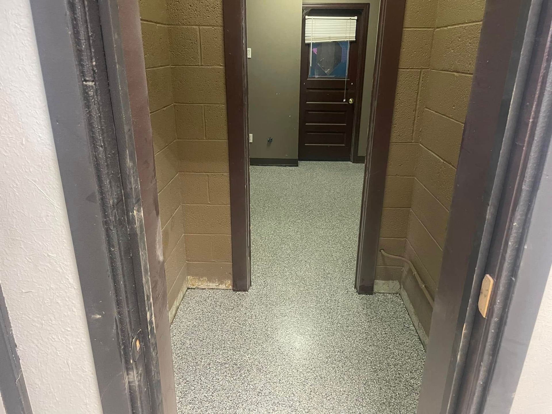Narrow hallway with speckled floor and two doorways. Dark door at end, window with blinds.