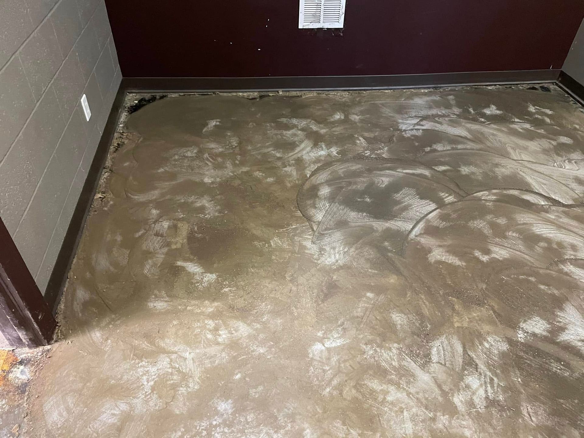 Floor with uneven, wet cement in a room with dark trim and beige/brown walls.
