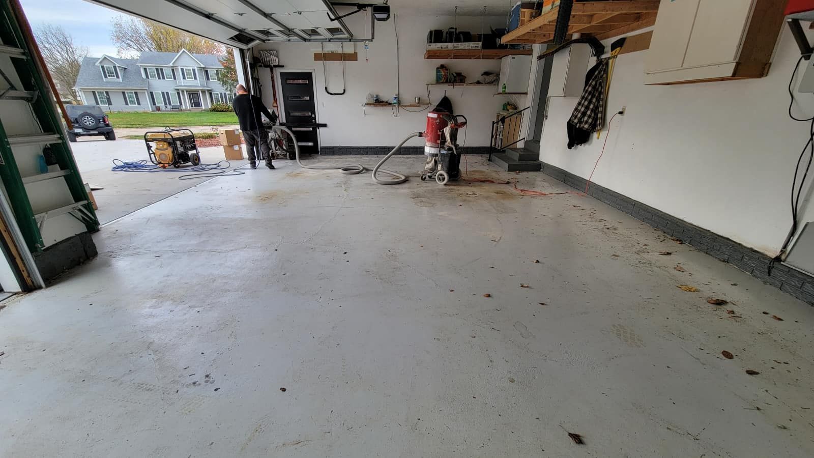 Person sweeping debris in a garage with another person present. Concrete floor, open garage door, natural light.