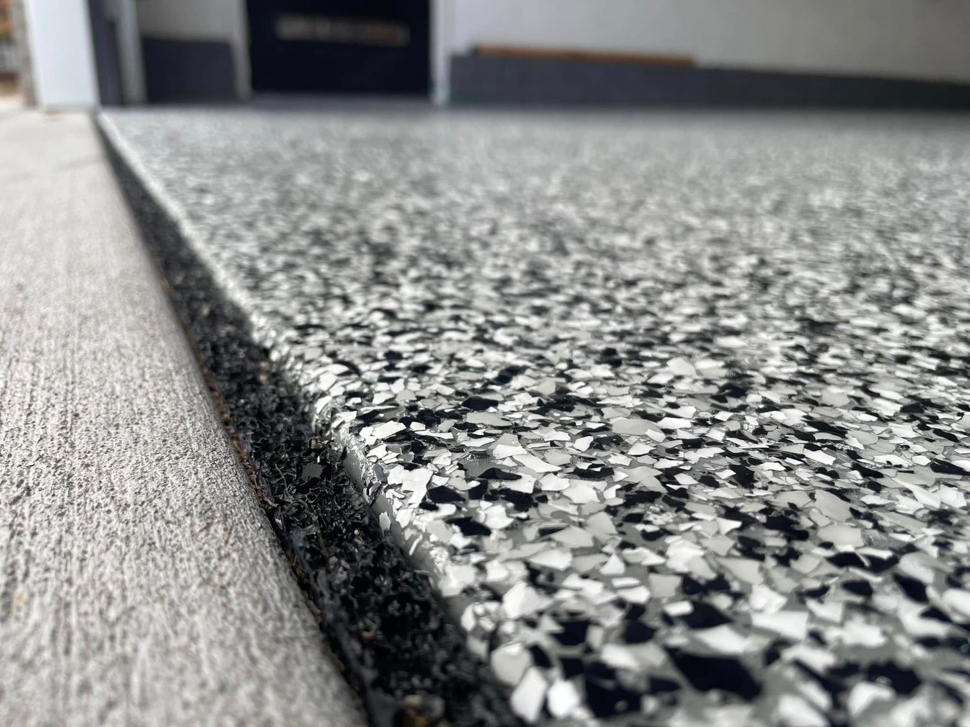 Close-up of a garage floor with a speckled epoxy coating, various shades of gray, bordering a concrete edge.