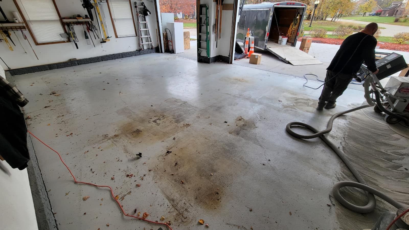 Man operating floor grinder in a garage. Gray epoxy floor, debris, open door with trailer visible.