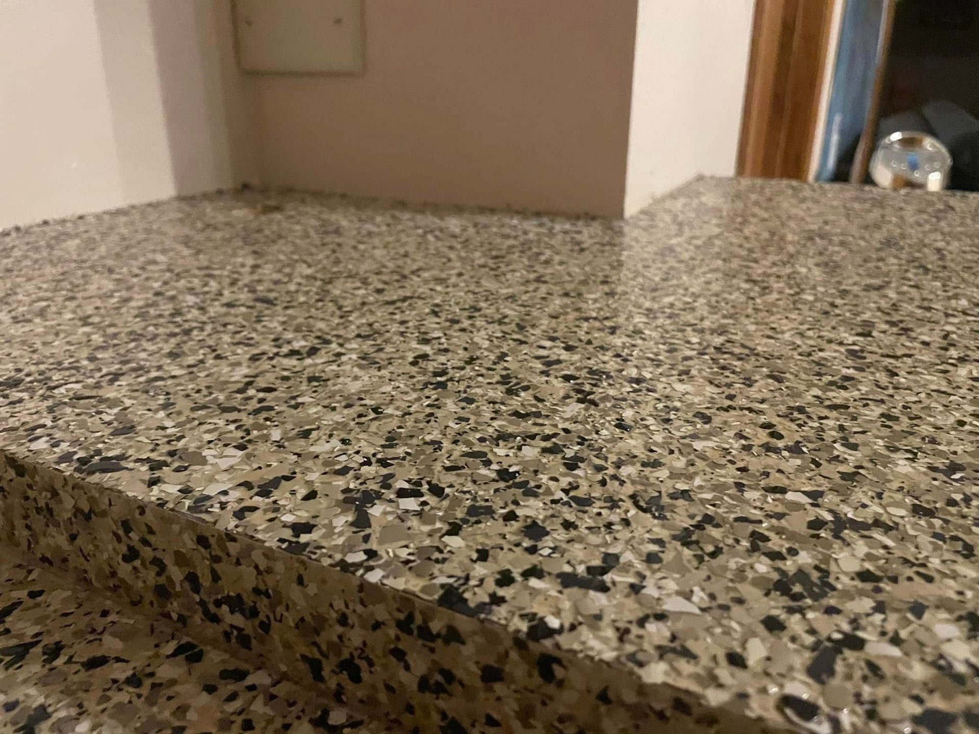 Close-up of a speckled countertop with beige, black, and gray chips; a light-colored wall is in the background.