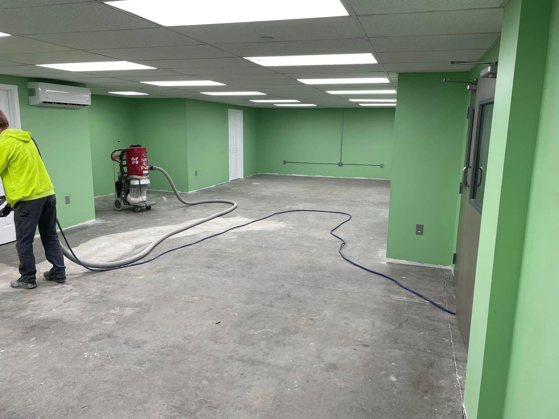 Person sanding concrete floor in a green-walled room. Black hose, red machine, overhead lights.