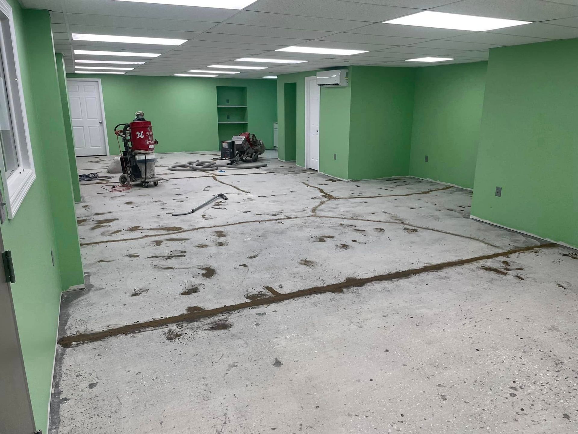 Room with green walls, concrete floor under construction; two workers, one using a grinder.