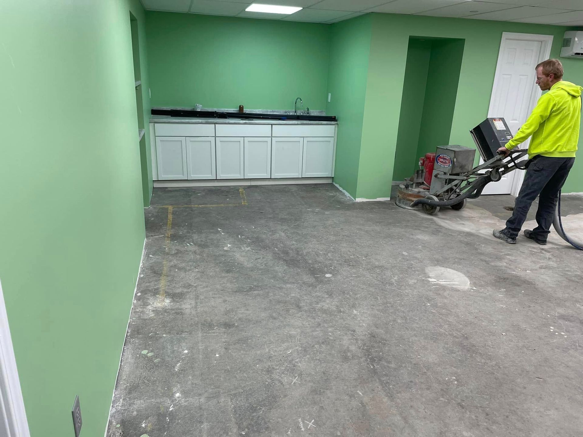 Man grinding a concrete floor in a room with green walls and white cabinets.