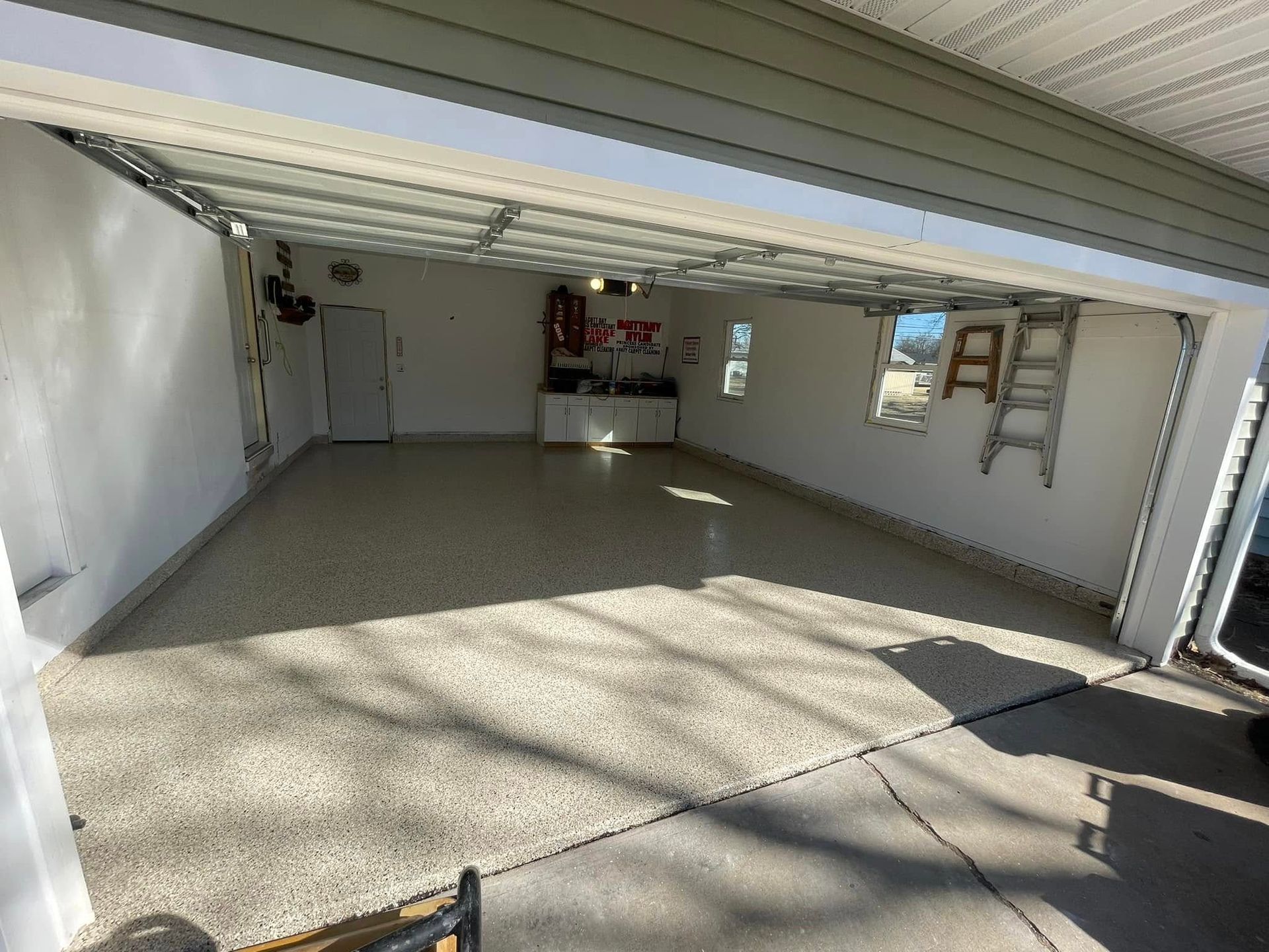 Empty garage with a speckled floor, open door, and a ladder leaning against the wall.