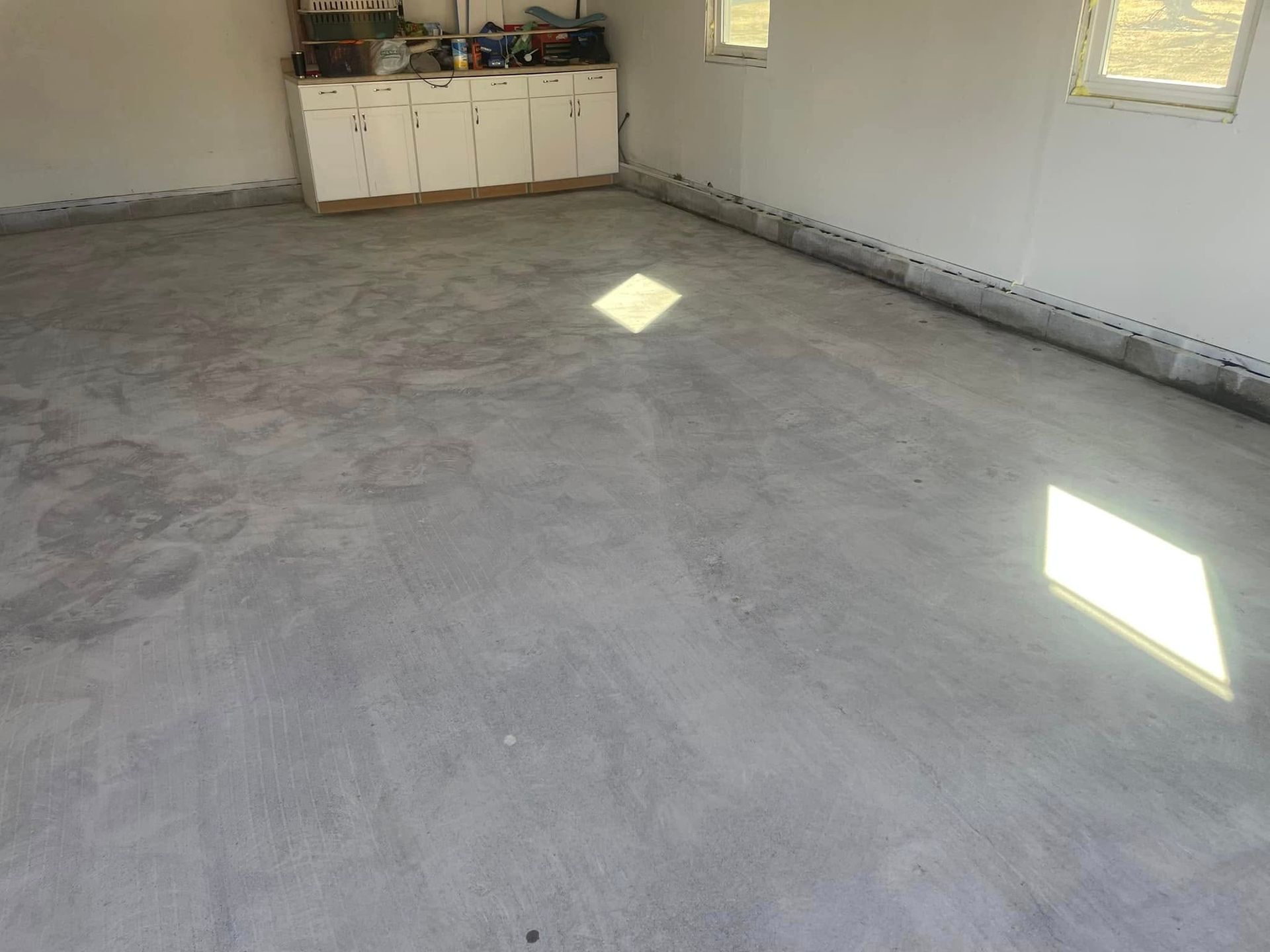 Garage interior with a gray concrete floor, white walls, and cabinets. Sunlight enters from windows.