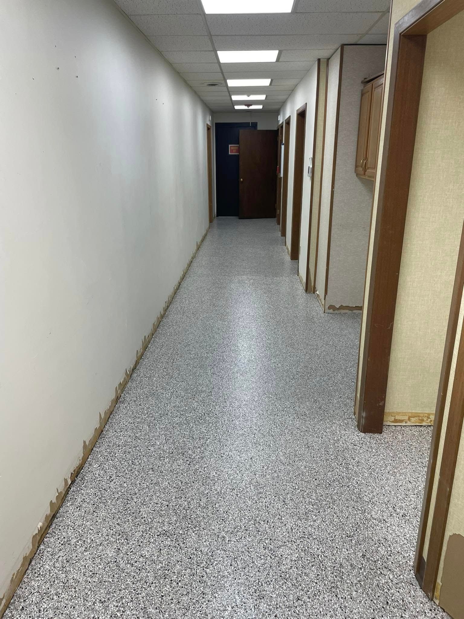 Long, narrow office hallway with speckled gray flooring, white wall, and brown door frames.