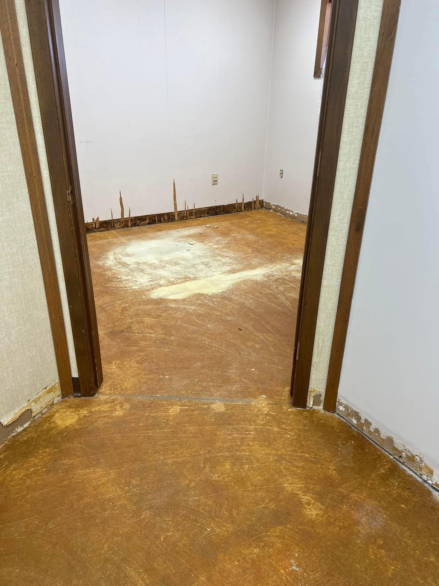 Room with brown floor covered in debris, doorway, white walls with brown trim.